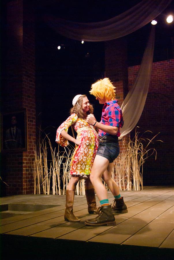 Two performers on stage, with one wearing a colorful dress and hat, and the other in a red and blue checkered shirt and shorts. Both wear boots, and are dancing energetically. The background has draped fabric and a rustic brick setting.