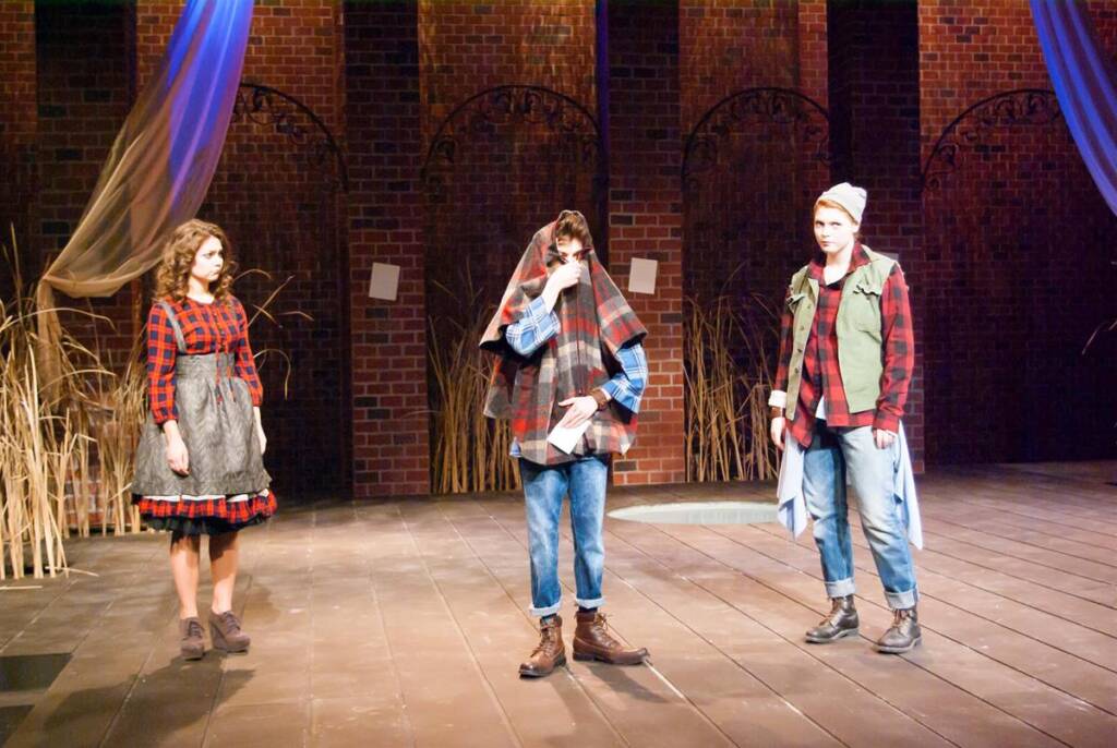 Three actors on stage in a play. The actor on the left wears a plaid dress and apron, the center actor is wrapped in a plaid blanket, and the right actor wears a beanie and layers. The background has brick arches and draped fabric.