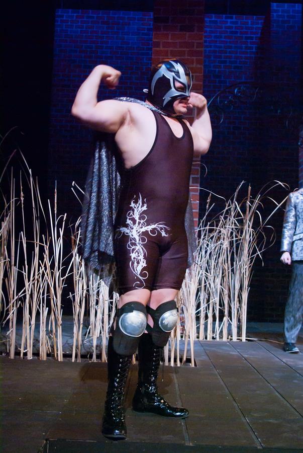 A wrestler in a black mask and decorated bodysuit flexes his arms on stage. He wears knee pads and boots, with tall reeds as part of the background decor. The setting is dimly lit with a brick wall behind him.