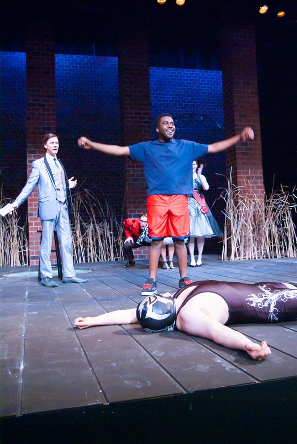 A man in a light suit gestures on a stage while another man in a blue shirt and red shorts stretches his arms triumphantly. A masked person lies on the floor in a wrestling pose. Two other people are in the background, with reeds as the backdrop.