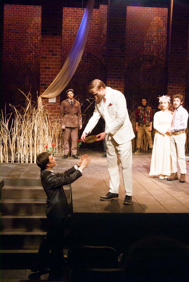 A theatrical performance with a man in a white suit standing on stage, extending a hand to another man in a black jacket below. Other actors in period costumes stand on a set with tall grasses and brick walls.