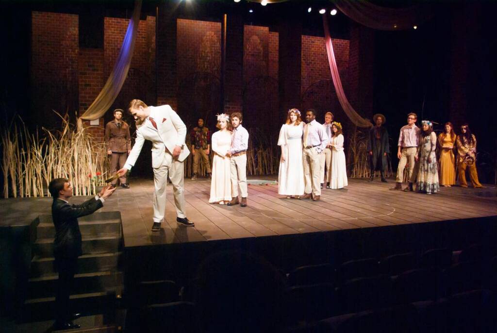 A man in a white suit on stage bends to accept a rose from a boy. Several people in period costumes stand nearby, with some dressed in white. Tall grass lines the brick backdrop. The stage is warmly lit.