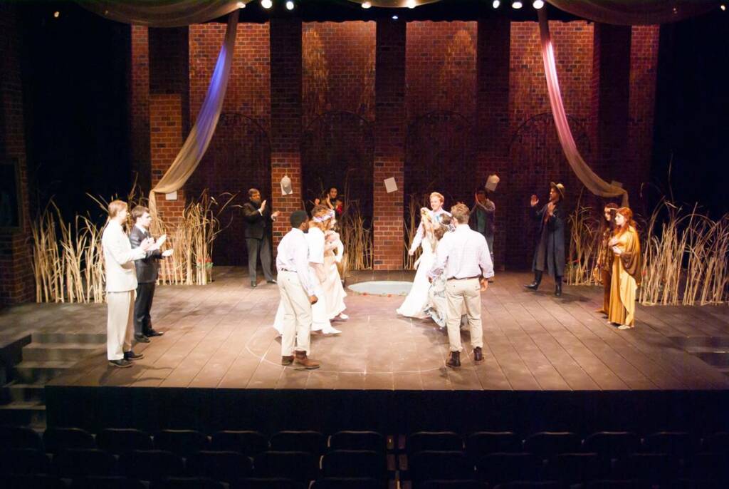 A stage performance with actors wearing period costumes. Some are clapping while others are engaged in a scene. Brick walls and decorative drapery set the backdrop, with tall grasses flanking the stage. Theater seats are visible in the foreground.