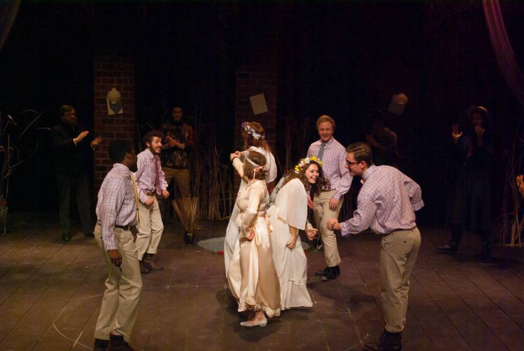 A group of people in costumes joyfully dances on a stage. They are dressed in various outfits, with some wearing floral crowns and long robes. The background features dim lighting and a brick wall.