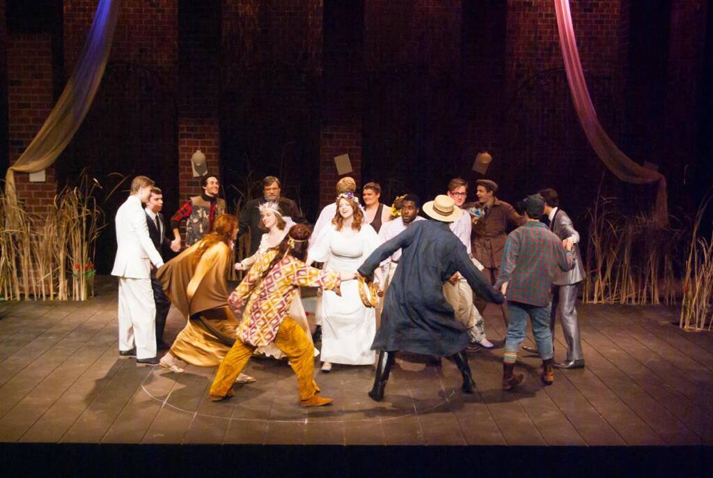 A group of performers in colorful costumes hold hands and form a circle on a stage. The scene is set against a backdrop of tall grass and hanging fabric. The atmosphere suggests a lively theatrical performance.