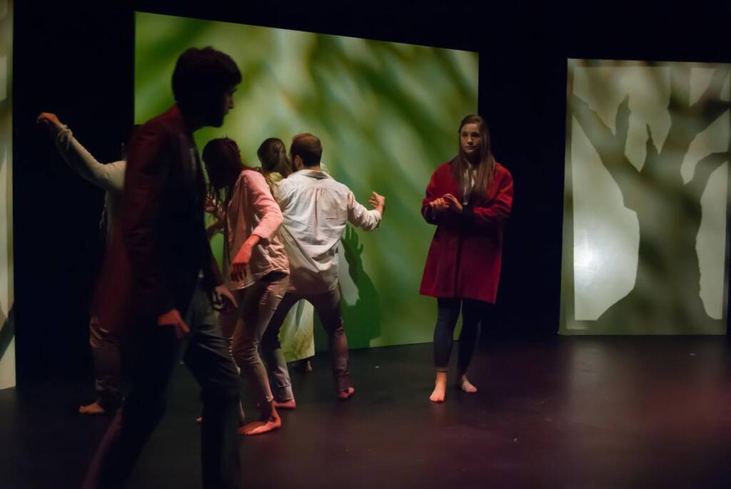 A group of people on stage, with one person in a red coat standing apart. The background features abstract, green leaf-like patterns on screens. The scene is dimly lit, creating a dramatic atmosphere. Some people are barefoot.