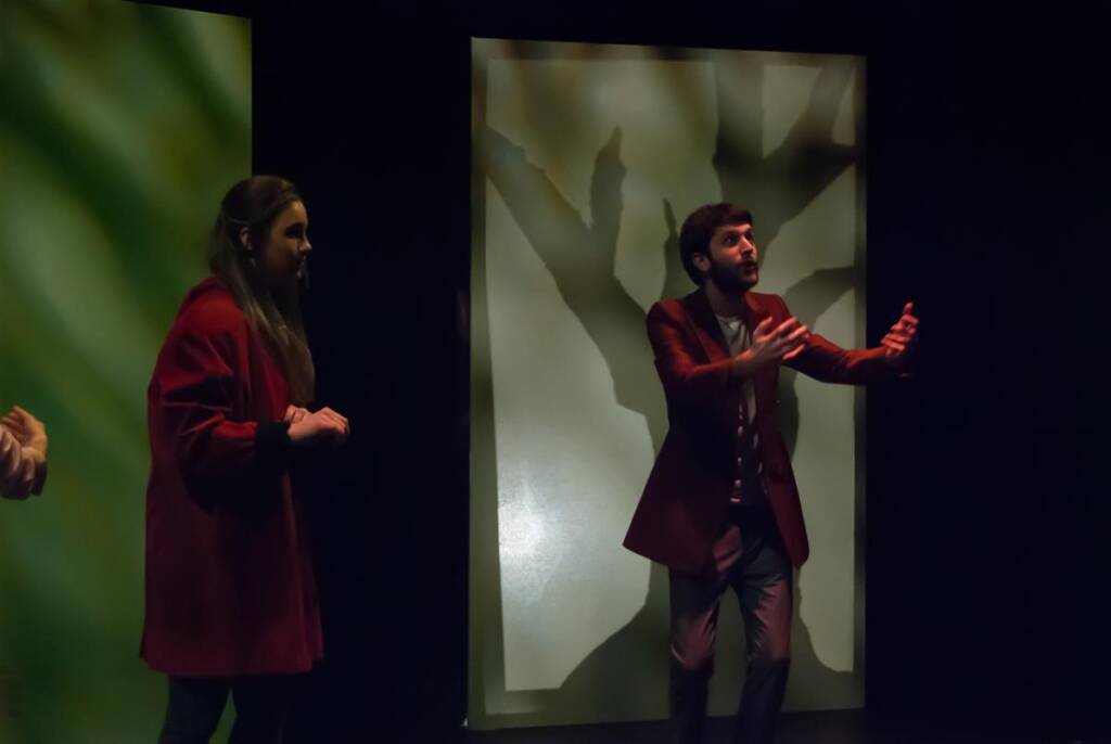 Two people on stage perform in dramatic lighting. The woman on the left wears a red coat and looks attentively at the man on the right, who gestures expressively with both hands. The backdrop features a shadowy, abstract tree pattern.