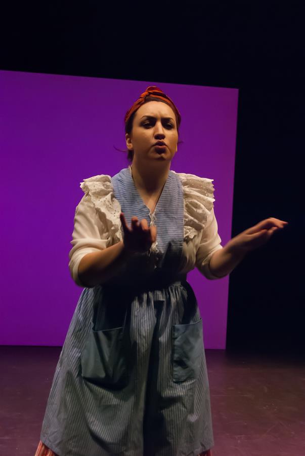 A woman with a red headscarf and blue striped apron gestures expressively on a dimly lit stage. She stands in front of a bright purple backdrop, wearing a blouse with ruffled sleeves.