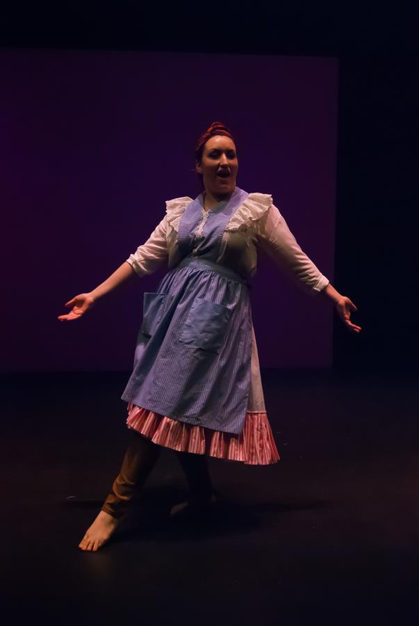 A performer on stage in a blue and white costume with a ruffled apron stands barefoot against a dark background. They have an expressive pose with arms extended, suggesting a theatrical performance or dance.