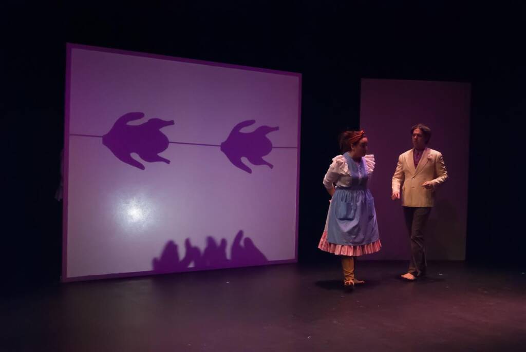 Two actors perform on stage. The woman wears a blue dress and apron, and the man wears a light-colored suit. Two shadow puppets resembling hands are projected on a screen behind them in a dark theater setting.