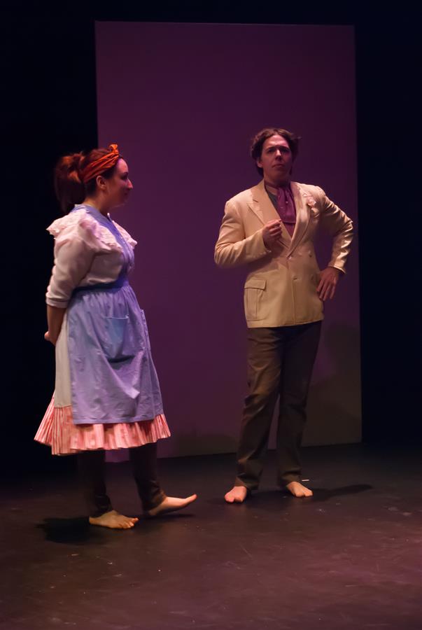 Two actors perform on stage. The woman on the left wears a blue apron and dress, with an orange headband. The man on the right is dressed in a beige suit and gesturing with one hand. Both are barefoot. The background is a dark, plain backdrop.