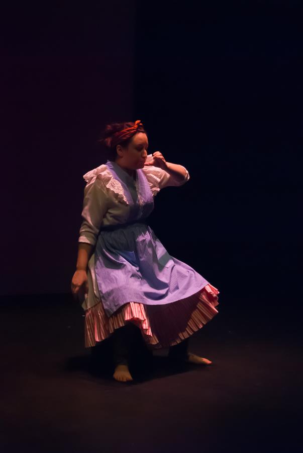 Person wearing a vintage-style dress with a blue apron and a headband, dancing on a dimly lit stage with a black background.