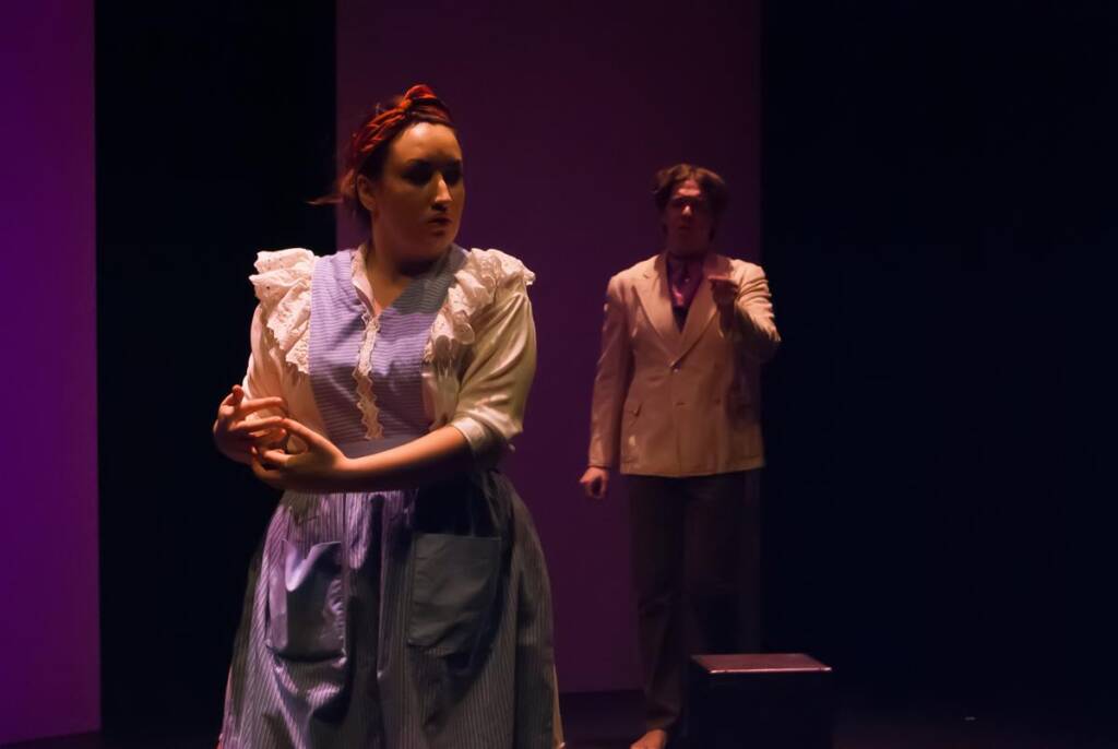 A woman in a striped dress with a ruffled apron stands in the foreground on stage, with her hands clasped. A person in a beige suit is in the background. The scene is dimly lit, suggesting a theatrical performance.