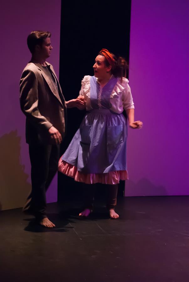 Two actors perform on stage. The man on the left wears a suit, and the woman on the right wears a long, blue dress with a pink underskirt and a red headband. The background features dark and purple lighting, creating a dramatic atmosphere.