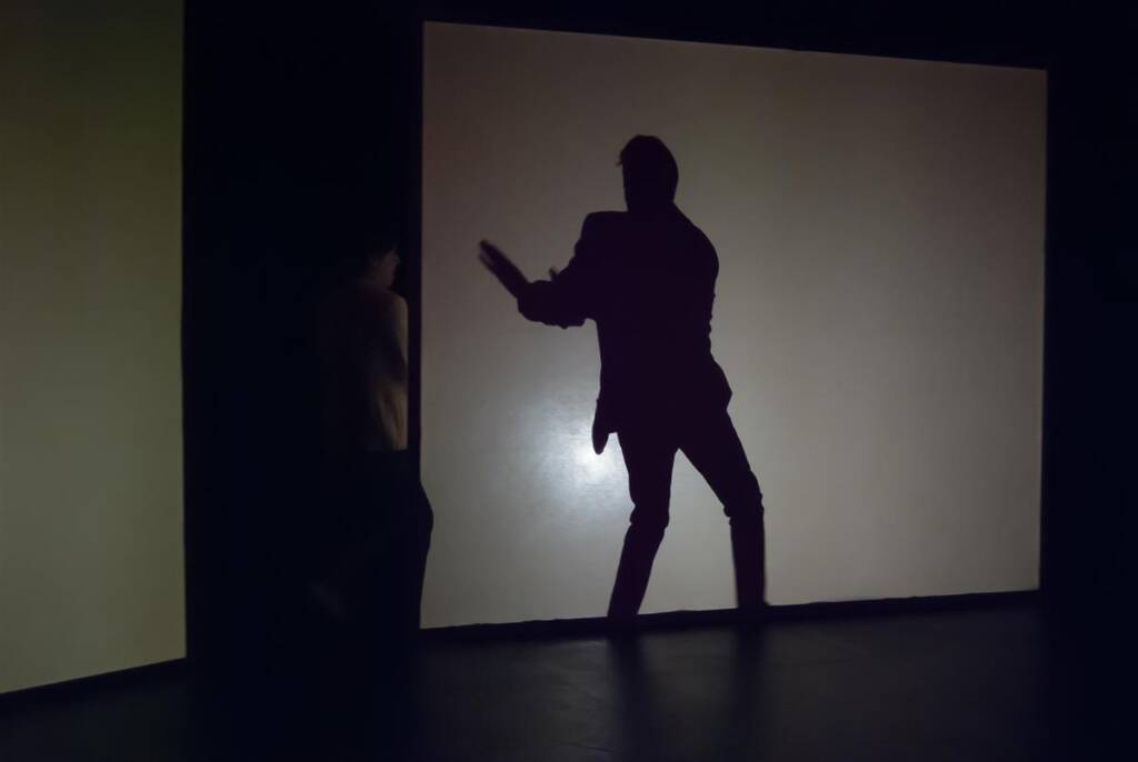 Silhouette of a person striking a playful pose against a bright backdrop, creating a sharp contrast. Another figure is partially visible on the left, emerging from the shadows. The setting suggests a creative or theatrical environment.