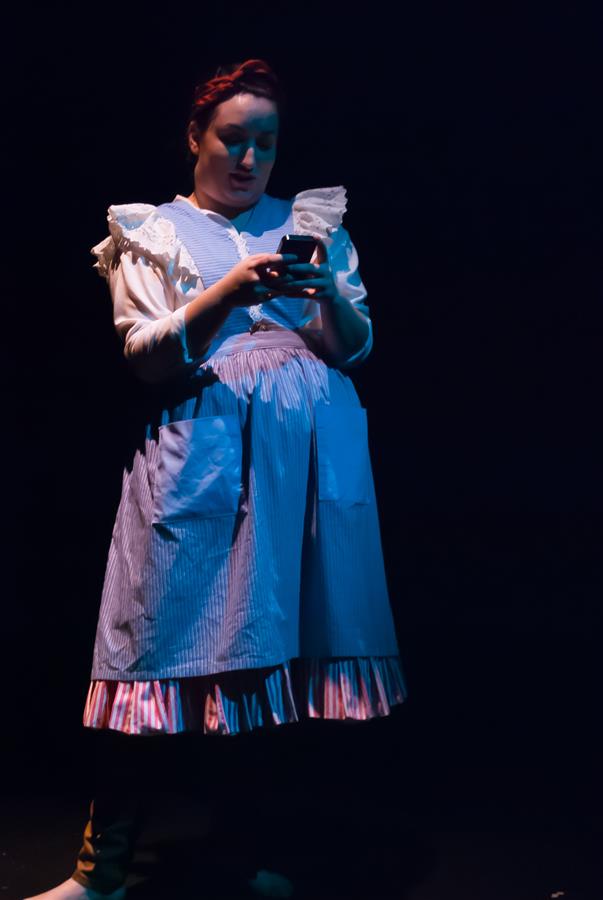 A person wearing a blue and white dress with striped patterns stands on a dimly lit stage, looking at a smartphone. The dress features ruffles and an apron-like front. The background is dark, creating a spotlight effect on the person.