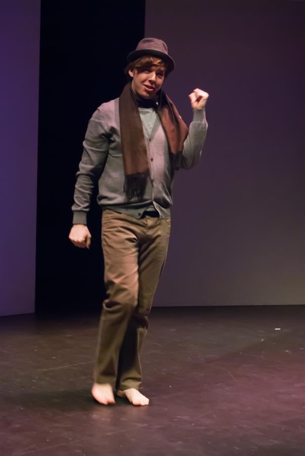 Person wearing a hat, scarf, sweater, and pants, performing barefoot on stage with one hand raised and the other by their side. The background is dark and the setting appears to be a theatrical performance.