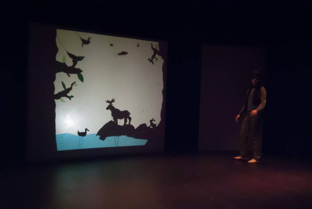 A person stands on a dark stage next to a large screen displaying a shadow puppet scene. The scene depicts various animals, including a stag and birds, against a backdrop of trees and a body of water.