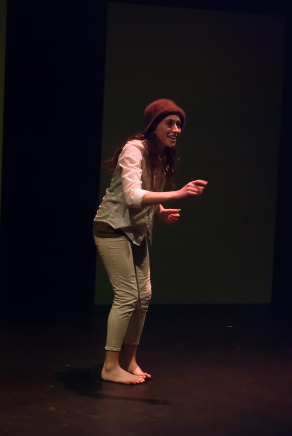 A person in casual clothing and a beanie performs on a dimly lit stage, smiling and gesturing expressively.