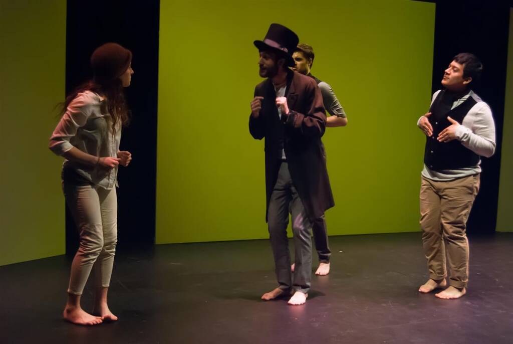 Four people are performing on stage. One wears a top hat and long coat, engaging with another in casual attire. Two others stand in the background, all barefoot. The backdrop is a plain, bright yellow-green screen.