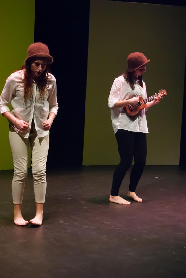 Two performers on stage both wearing brown hats and white shirts. The person on the left is walking with a serious expression, while the person on the right is playing a ukulele. The background is green and black.