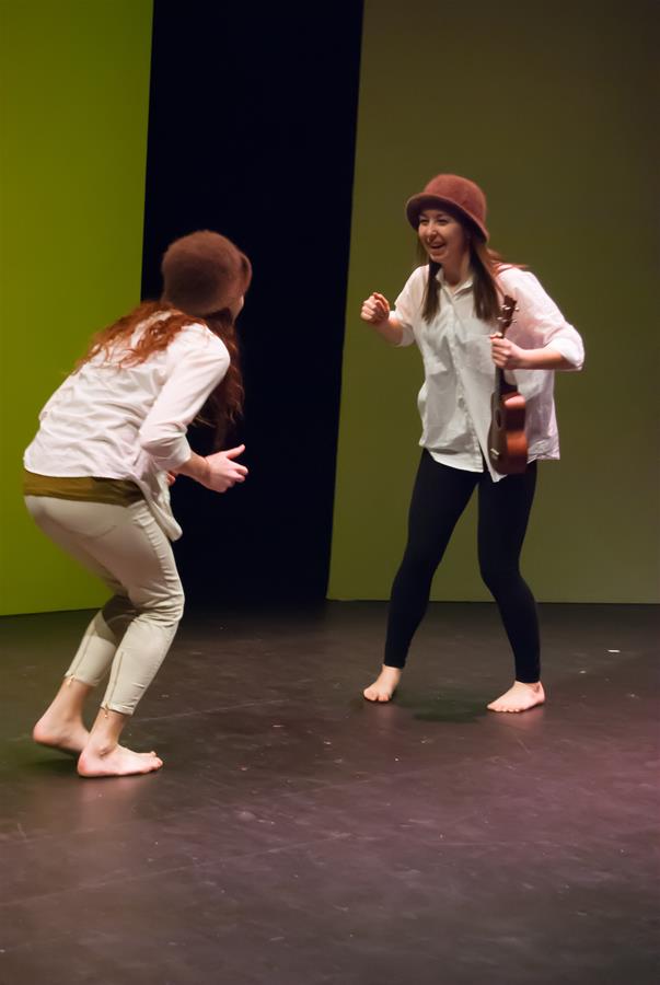 Two barefoot women perform on stage, one with a ukulele and a brown hat, both smiling and dressed casually. They're in a dynamic pose, suggesting playfulness or a skit. The backdrop is divided into green and dark sections.