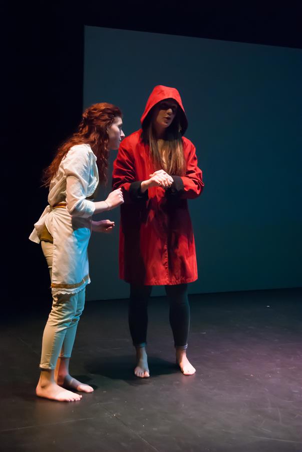 Two actors perform on stage. One wears a white outfit, while the other wears a red hooded coat. Both are barefoot and engaged in an expressive scene against a dark backdrop.