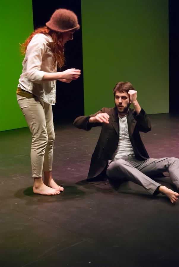 Two people performing on stage. A barefoot woman in light pants and a hat leans slightly over a seated man with a beard, who gestures energetically. The background is lit in green.