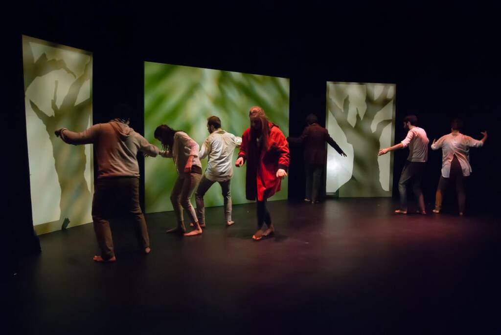 A group of people on stage are performing in low light. Six individuals are spread out, facing large screens with nature-inspired patterns. The setting is dramatic with soft spotlighting highlighting their poses and movements.