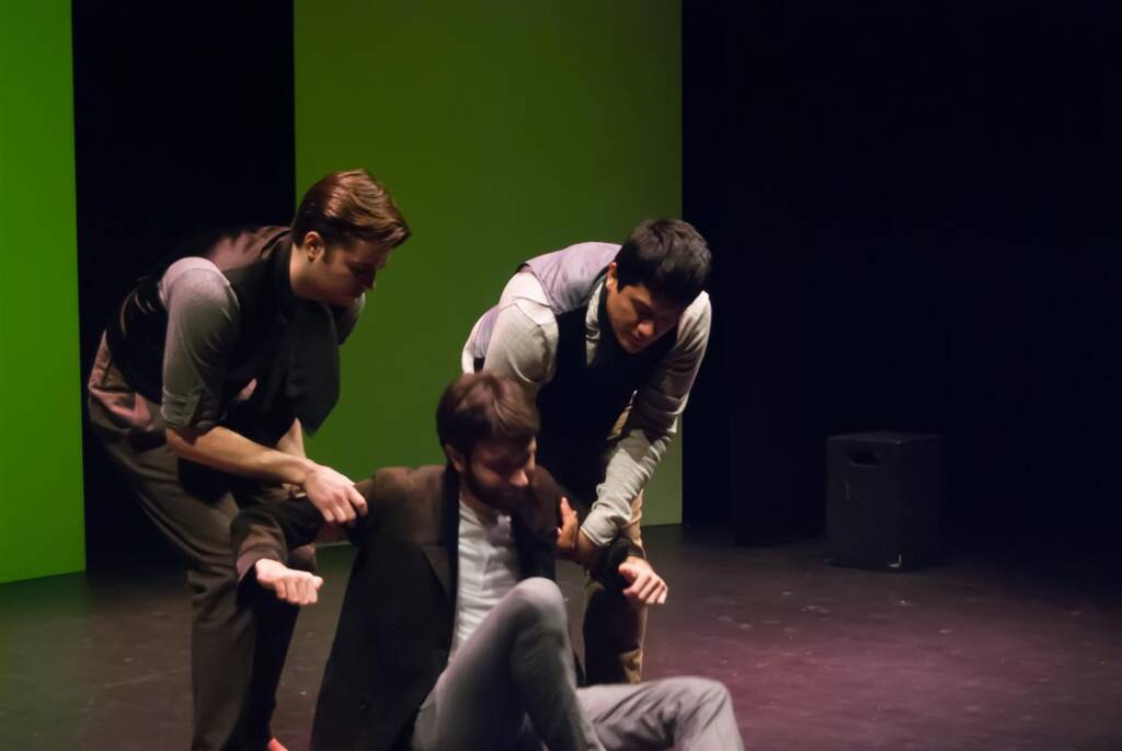 Three actors are performing on stage. Two are standing and helping a third actor, who is sitting on the floor, to stand up. They are dressed in formal attire against a backdrop with a bright green and black color scheme.