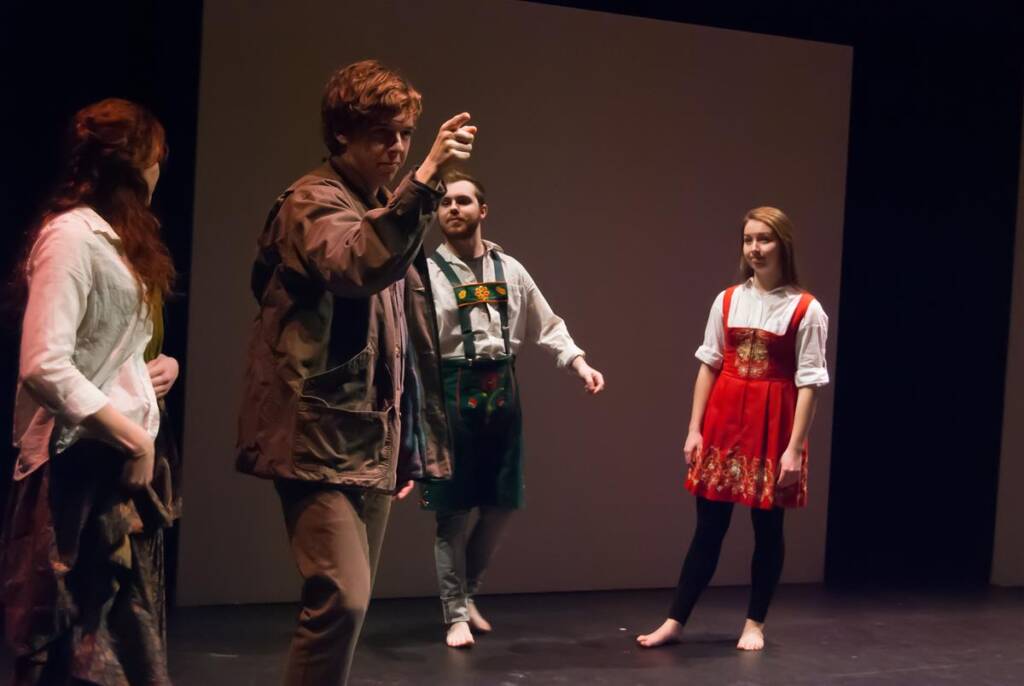 Four actors perform on stage. One gestures dramatically, wearing a jacket and pants. The others are dressed in traditional attire, with two women in dresses and a man in a vest and shorts. The background is simple and dark.