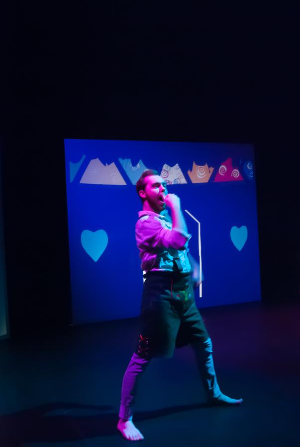 A barefoot person in expressive pose on stage, wearing a white shirt and dark pants. The backdrop is blue with pink and white graphics, including hearts and abstract shapes. The scene is dimly lit with a spotlight on the performer.