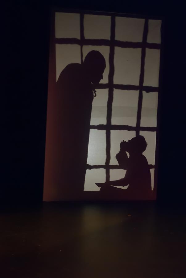 Silhouetted image of a tall person standing and a seated person in front of a window with a grid pattern. The scene is dimly lit, casting shadows on a backdrop.