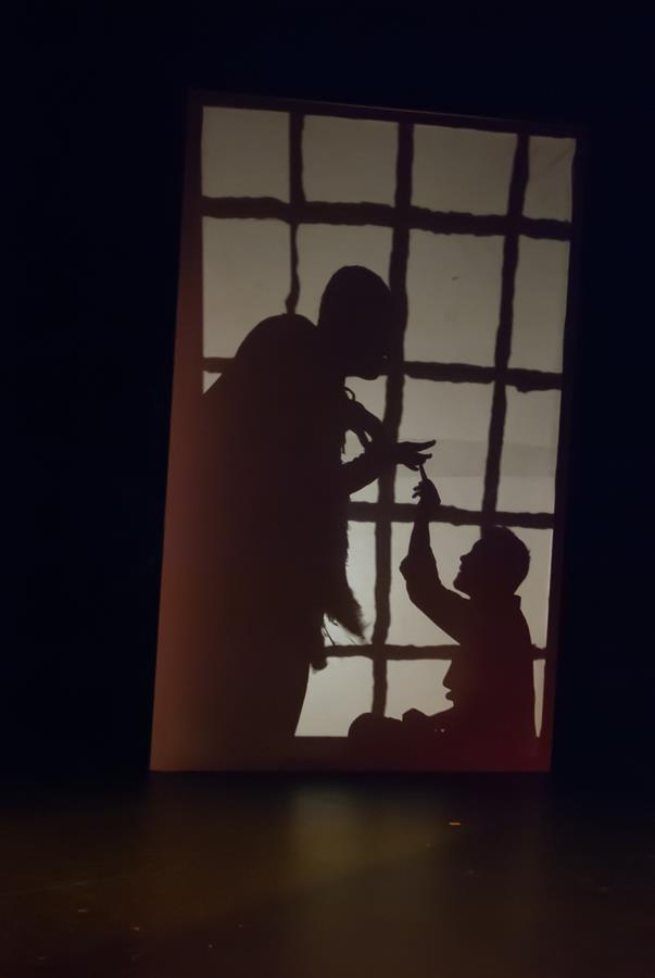 Silhouettes of two people are seen against a backlit, gridded screen. One is standing, pointing downward, while the other is kneeling, gesturing upward, creating a dramatic scene on stage.