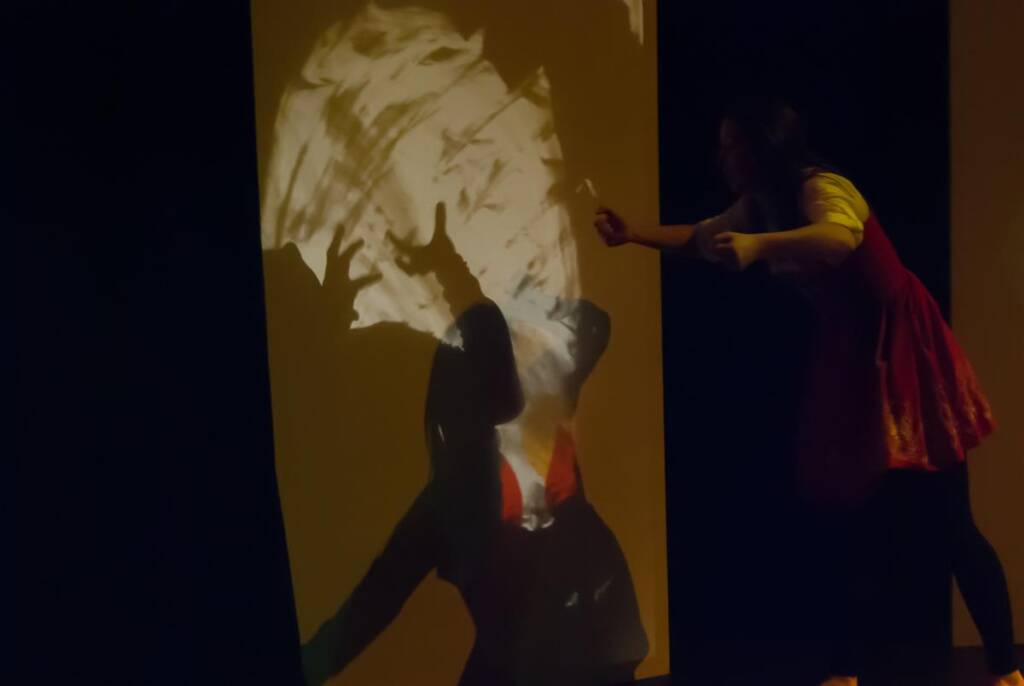 A person on stage interacts with a shadow projected onto a large screen. The shadow depicts an exaggerated figure in dramatic light, as the person reaches out, creating an artistic and mysterious atmosphere.