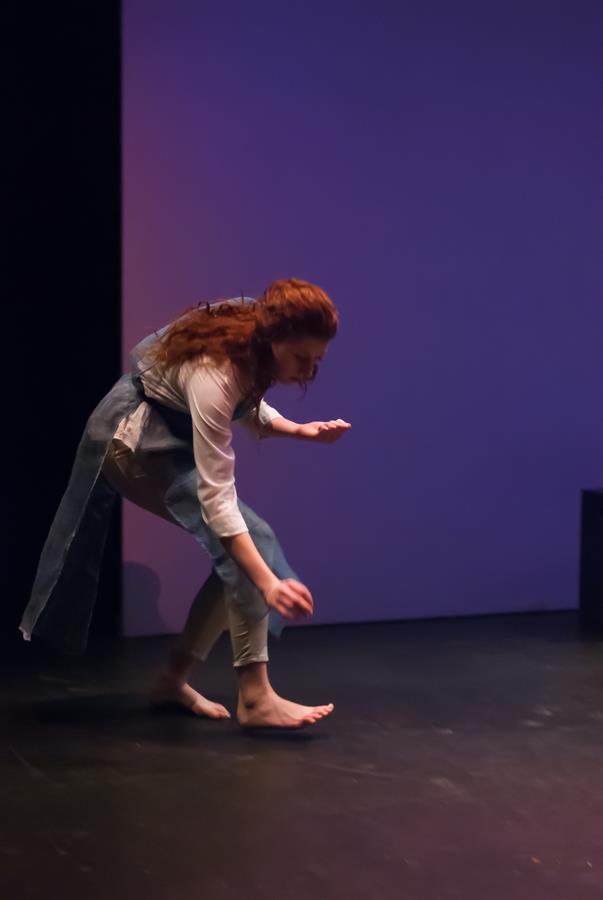 A barefoot person with long red hair is performing a dance on stage, bent forward with one arm extended down and the other raised. They wear a white shirt and blue garment. The background is a muted blue with dramatic lighting.