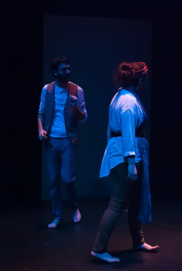 Two actors perform on a dimly lit stage. A man with a beard, wearing a vest and pants, gestures with one hand. A woman with a headband, wearing a white top and sash, stands with her back to him. Both are barefoot, creating a dramatic atmosphere.