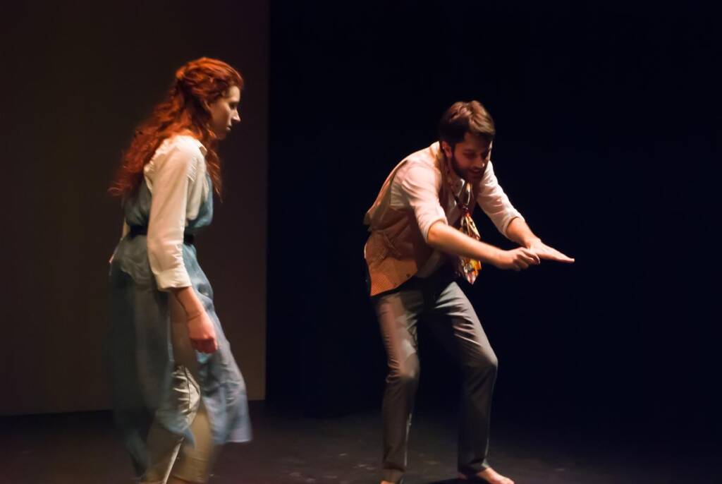 Two actors perform on stage. One, a woman with long red hair in a blue and white dress, stands on the left. The other, a man with a beard wearing a vest, gestures animatedly with his arms extended forward. The background is dark.