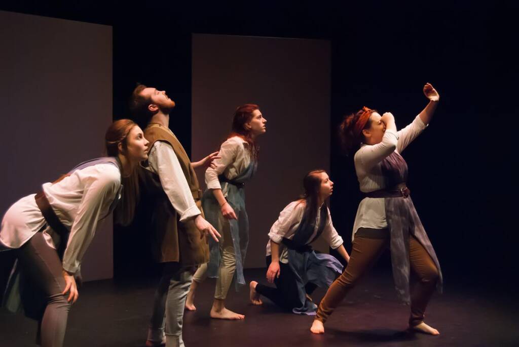 Five actors on stage in dramatic poses, facing left. They wear historical or fantasy-style costumes with tunics and belts. The lighting is dark and focused, creating an intense atmosphere. The background is plain, emphasizing the performers' actions.