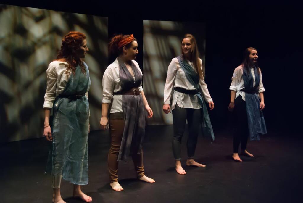 Four people stand barefoot on stage, engaged in conversation. They are dressed in casual attire with fabric sashes over their shoulders. The background is dark, with patterned lighting creating abstract designs on the wall.