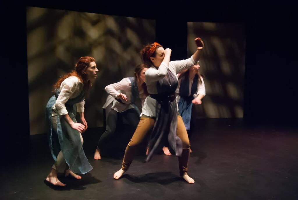 Four performers on stage enact a dynamic scene. One person is in a dramatic pose, while the others watch attentively. The background is dark with patterned lighting, creating an intense atmosphere. All are dressed in casual, period-style clothing.