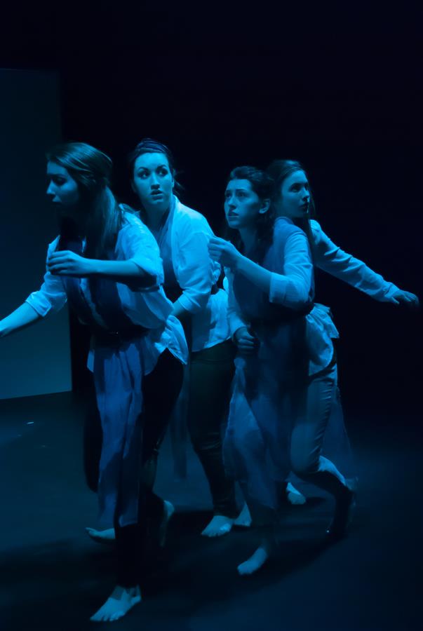 Four people stand closely together on a dimly lit stage, illuminated by blue light. They appear focused and intense, with some looking forward and others to the side, all barefoot and wearing casual clothes.