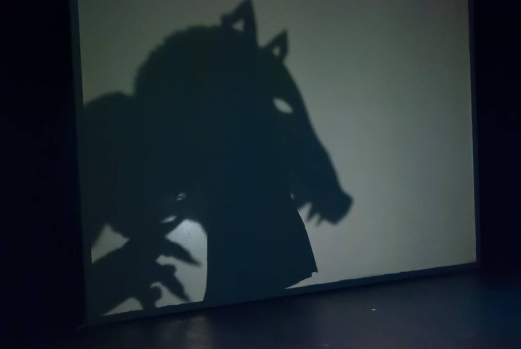 Shadow of a person creating a wolf-like shape with their hands and head, projected on a wall in front of a dark background.