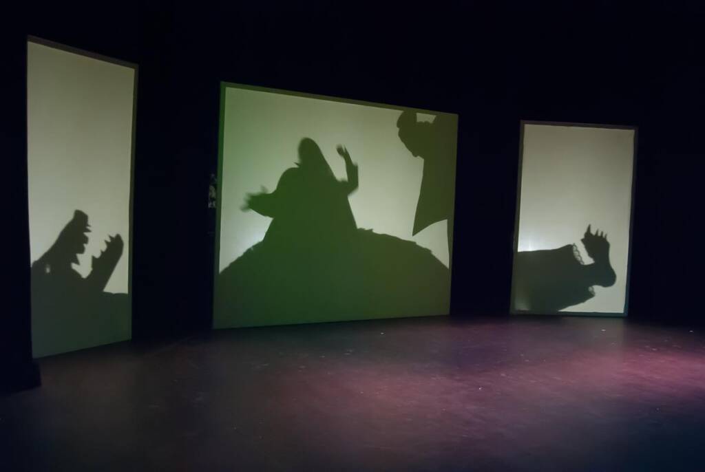 Silhouettes of dinosaur and human figures are projected onto three vertical screens in a dark room, creating a dramatic shadow effect. The scene appears theatrical, with dynamic poses suggesting interaction and movement.