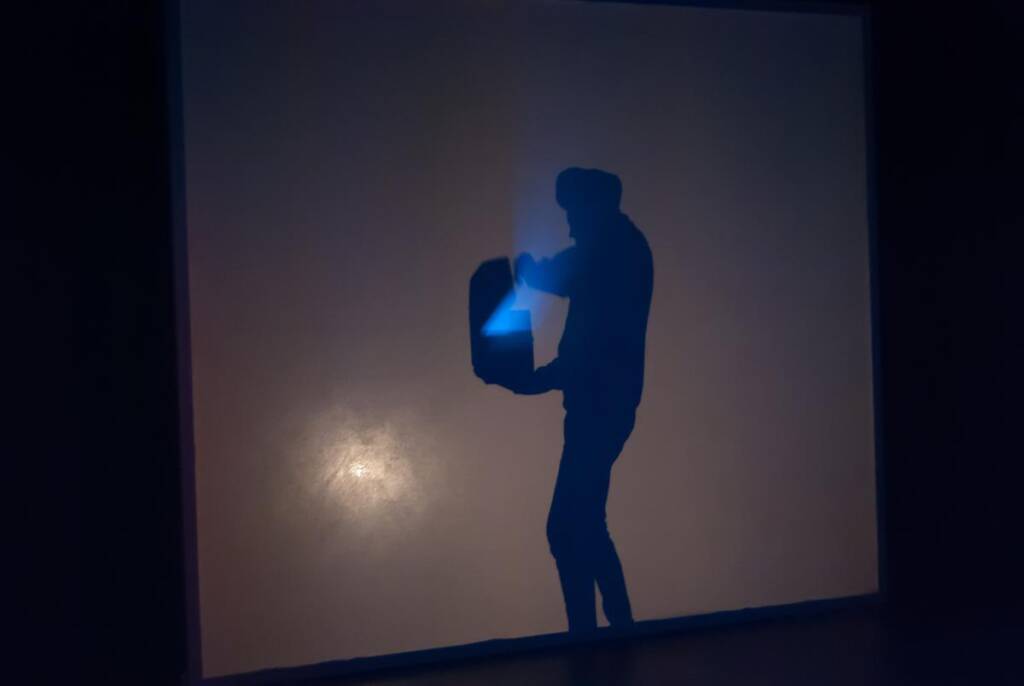 A silhouette of a person with a hat stands against a dimly lit background, holding an open suitcase that emits a blue light.