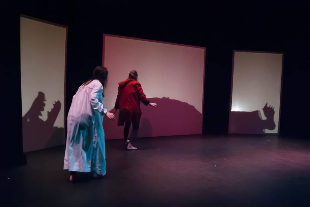 Two people onstage, one in a white outfit and the other in red, are interacting with dinosaur shadows projected on screens behind them. The stage is dimly lit, creating a dramatic ambiance.