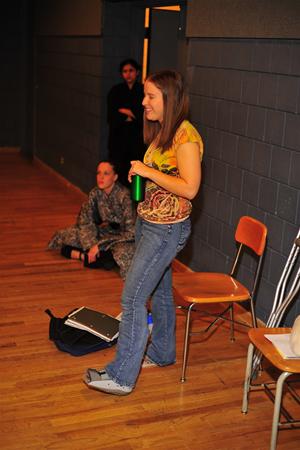 A woman in a yellow patterned shirt and jeans stands holding a green bottle, smiling. Behind her, two people sit on the floor near a wall, one in a camouflage uniform. An empty wooden chair and some papers are nearby.