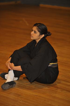 A person wearing a black martial arts uniform sits on a wooden floor with legs crossed and arms wrapped around their knees. They have dark hair tied back and are looking intently to the side.