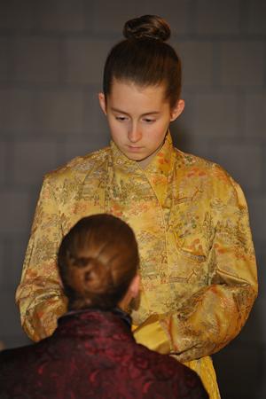 A person with long brown hair in a bun is wearing a gold, intricately patterned robe, looking down at another person with similar hair in a bun, wearing a dark red robe. The background is a neutral-colored wall.