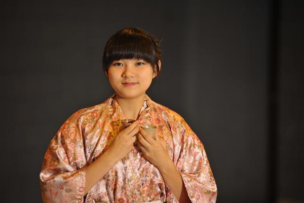 A person wearing a floral-patterned kimono holds a small cup with both hands. The background is dark and blurred, emphasizing the person.